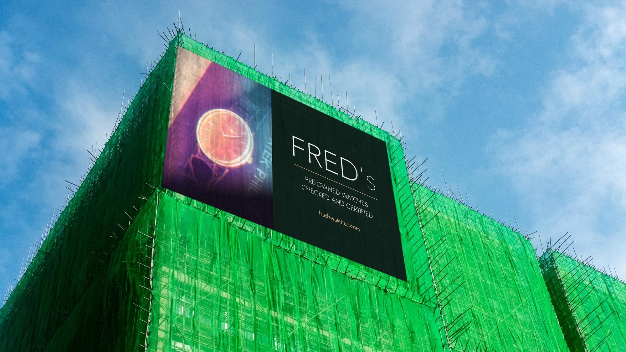 A building wrapped in green scaffolding net with a large FRED's billboard showing a gold watch on a strap